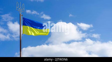 Ukraine blue yellow flag waving over blue sky with white clouds on flagpole with Coat of Arms of Ukraine State emblem National ukrainian symbol Triden Stock Photo