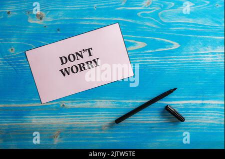 Light pink paper with Dont worry sign on it placed over textured blue wooden background with black marker beside it. Stock Photo
