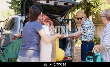 Big family packing voyage luggage in car trunk while getting ready for summer field trip. Happy people loading vehicle with luggage and trolleys while going on weekend citybreak together. Stock Photo