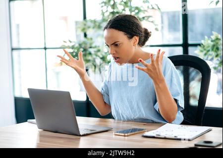 Irritated angry young mixed race business lady, company director, supervisor, annoyed looking at laptop screen while sitting in office, gesturing with hands, annoyed by poor performance of employees Stock Photo