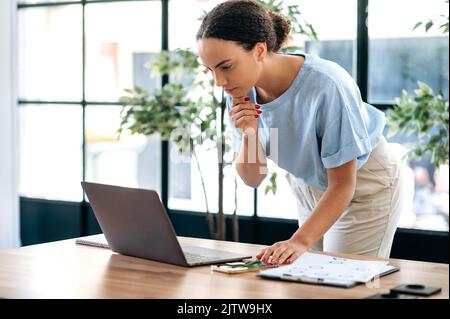 Focused busy brazilian or hispanic female employee, talking on smartphone during work paperwork project, stand in modern office. Successful mixed race business woman having cell phone conversation Stock Photo