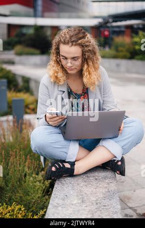 Woman student with curly hair and eyeglasses using laptop computer, mobile phone, studying, learning language. Communication, internet concept Stock Photo