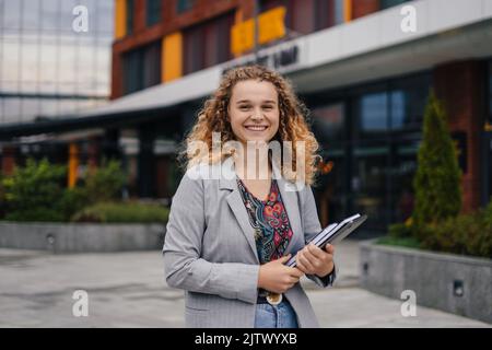 Beautiful smiling caucasian student holding books and laptop, looking at camera walking on university campus. Having break in classes. Urban and Stock Photo