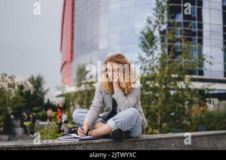 Pensive woman taking notes, writing resume sitting outside the university building. Taking notes learning college academic educational course concept Stock Photo