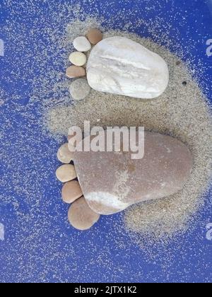 Baby feet shape made with pebbles on the beach with sand and blue background. Vertical banner Stock Photo