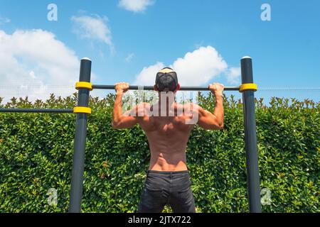Muscular man in a cap doing pull-ups on the horizontal bar in the park outdoors, back view of his back Stock Photo