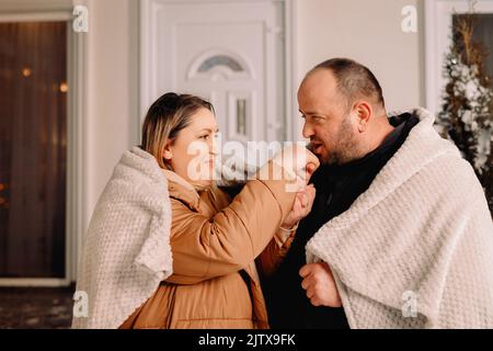 Close up photo of woman and man standing in front the house on evening wearin warm clothes covered wirh blanket and man warms wife hands with his Stock Photo