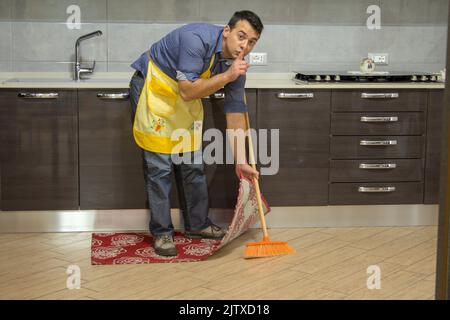 boy doing housework while making a gesture of silence and hiding dirt under the carpet Stock Photo