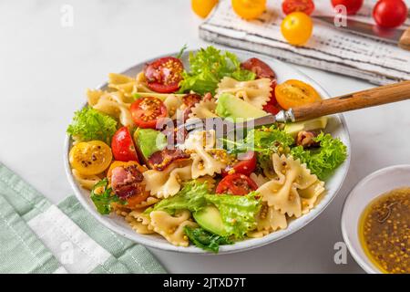 Cold summer pasta salad with bacon, tomatoes, avocado and mustard in a plate with fork on white background. Healthy diet food Stock Photo
