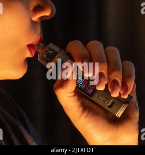 A girl smokes an electronic cigarette and exhales smoke, her lips are painted with red lipstick, a portrait of a smoking girl on a black background. Stock Photo