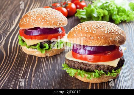 Two homemade cheeseburgers with beef patties , fresh salad, tomatoes and onion on seasame buns, served on brown wooden table. Stock Photo