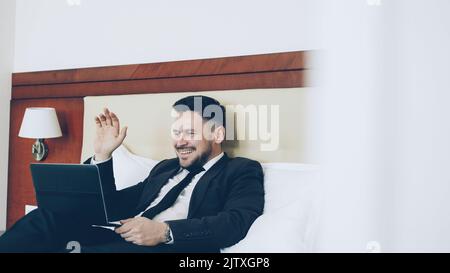 Happy cheerful businessman talking with family via online video chat using laptop computer lying on bed in hotel room. Travel, business and people concept Stock Photo