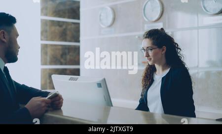 Receptionist girl talking with bearded businessman about check-in and giving key card to man at reception in hotel. Business, travel and people concept Stock Photo