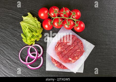 The raw ingredients for the homemade burger. Two raw coutlet on paper, tomatoes, salad, onion rings on black slate background. Top view. Flat lay. Stock Photo