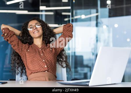 Young woman engineer developer programmer works in the office inside the IT company, satisfied with the work result, rests hispanic looks out the window, smiles hands behind her head, dreams. Stock Photo