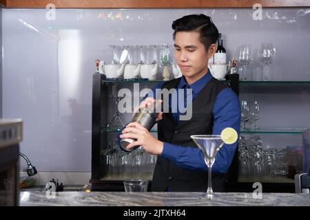 Young Asian barman using shaker to mix ingredients for the cocktails Stock Photo