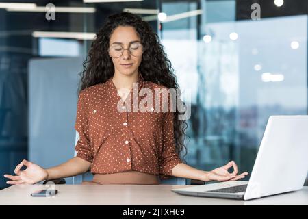Young beautiful Hispanic web developer working inside office building, woman resting meditating at workplace with eyes closed, female worker using laptop to write code. Stock Photo