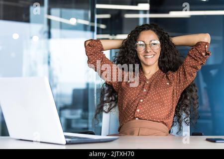 Young woman engineer developer programmer works in the office inside the IT company, satisfied with the work result, rests hispanic looks out the window, smiles hands behind her head, dreams. Stock Photo