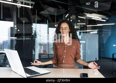 Young beautiful Hispanic web developer working inside office building, woman resting meditating at workplace with eyes closed, female worker using laptop to write code. Stock Photo