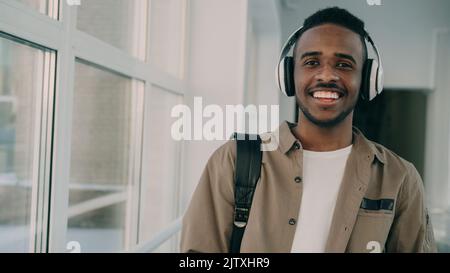Portrait of young handsome student of african-american ethnicity standing in wide white spacious corridor of college with big headphones on his neck looking at camera and smiling. Stock Photo