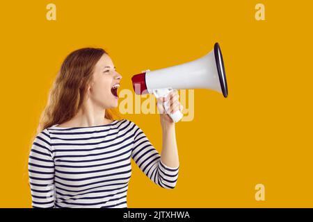 Young woman wearing striped jersey is shouting in loudspeaker on yellow background. Stock Photo