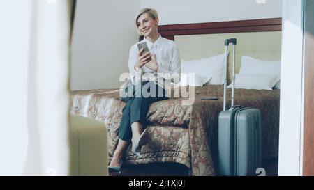 Happy cheerful businesswoman talking with family via online video chat using smrtphone camera sitting on bed in hotel room. Travel, business and people concept Stock Photo