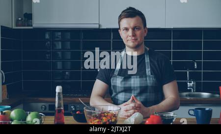 Portrait of young handsome caucasian executive chef in apron sitting at table in modern lighty spacious kitchen in front of plates , he is smiling, calm and positive, looking at camera Stock Photo