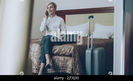 PAN of smiling blonde businesswoman in white shirt using on laptop and talking at mobile phone while sitting on bed in hotel room. Business, travel and people concept Stock Photo