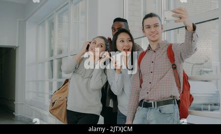 Group of four multi-ethnic students taking selfie on smartphone camera while standing in corridor of university . Hipster guy holding phone and friends are posing positively Stock Photo