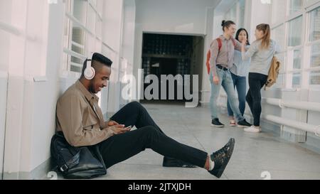 Young handsome african american male student sitting on floor in white corridor with headphones on head listening to music holding smartphone texting someone. His classmates are behind him Stock Photo