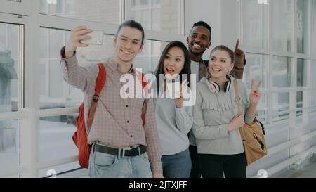 Group of four multi-ethnic positive male and female students are standing in wide white corridor of college. Hipster guy is holding smartphone making selfie of them all Stock Photo