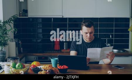 Worried young man paying bills online using laptop in the kithcen at home indoors Stock Photo