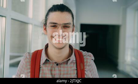 Portrait photo of young handsome attractive male student of caucasian ethnicity wearing glasses standing in wide white hallway indoors looking at camera and smiling positively Stock Photo