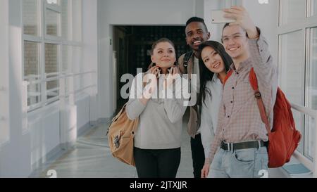 Group of four multi-ethnic students taking selfie on smartphone camera while standing in corridor of university . Hipster guy holding phone and friends are posing positively Stock Photo