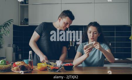 Attractive couple in the kitchen at home. Curly girl playing video game on smartphone while her boylfriend cooking breakfast Stock Photo