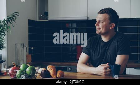 Portrait of young happy caucasian man sitting at table in modern spacious kitchen at home, looking through window, holding cup drinking tea, he is positive and cheerful Stock Photo