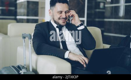 Tilt up of confident attractive businessman talking mobile phone and using laptop computer while sitting on armchair in luxury hotel with luggage suitcase near him. Travel, business and people concept Stock Photo