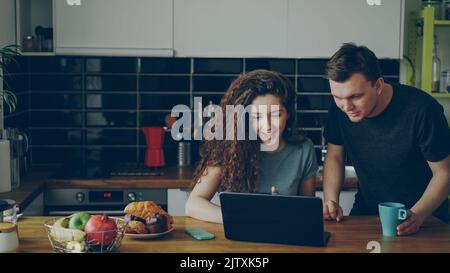 Young positive caucasian couple sitting at table in kitchen using laptop smiling and discussing at home Stock Photo