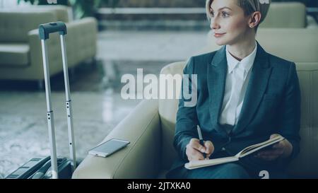 Attractive blonde businesswoman sitting in armchair in hotel lobby and writing in notepad smiling and looking aside. Business, travel and people concept Stock Photo