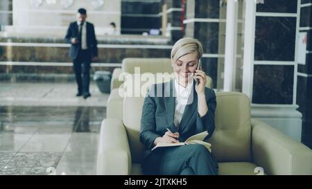 Attractive businesswoman sitting in armchair with notepad and laptop computer while businessman with luggage walking through hotel lobby from reception desk Stock Photo