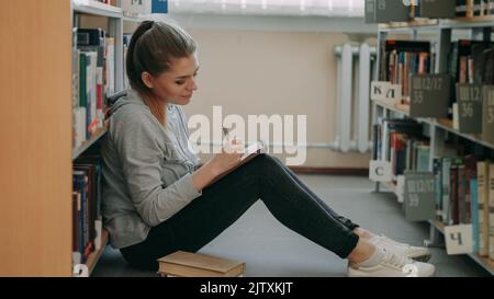 Young beautiful dreamy caucasian female student is sitting on floor in big lighty library among bookshelves writing down composition in copybook smiling looking up at ceiling. Stock Photo