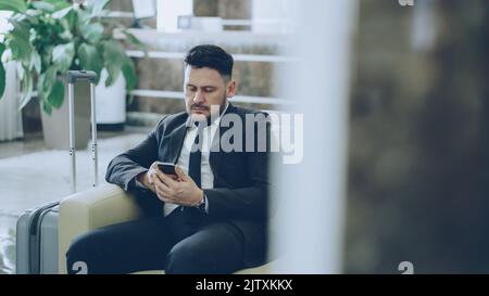 Bearded concentrated businessman using smartphone sitting on armchair inside luxury hotel after arrivel to business meeting Stock Photo