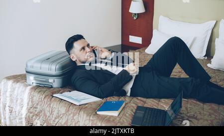 PAN of confident young businessman talking mobile phone while lying on bed in hotel room . Travel, business and people concept Stock Photo