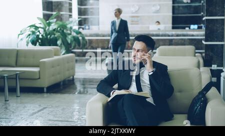 Pan shot of handsome bearded businessman sitting in armchair talking mobile phone with notepad while businesswoman in suit with luggage walking through hotel lobby from reception desk. Stock Photo