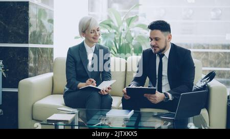 Two female and male business colleagues with clipboard and notepad having conversation at hotel lobby. Luggage in near couch. Business, travel and people concept Stock Photo