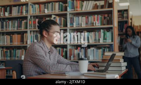 Young handsome hipster male student is sitting at table in lighty big spacious library with modern laptop Stock Photo