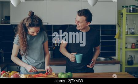 Curly woman cooking near table cutting red pepper in kitchen at home, her boyfriend showing her something funny in smartphone and they are smiling Stock Photo