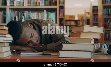 African american handsome young male student is sitting at table with big piles of books in light library. His head is lieing on table he is sleepingwith his hands under head Stock Photo