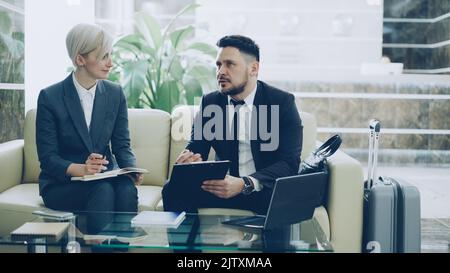 Confident businessman with clipboard talking to female colleague with notepad in hotel lobby prepraing for business meeting they arrived to. Luggage is near couch Stock Photo