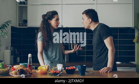 Pretty woman standing at table cutting salad while her boyfriend comes and they argueing speaking loud and nervous. Relations , difficulties concept Stock Photo